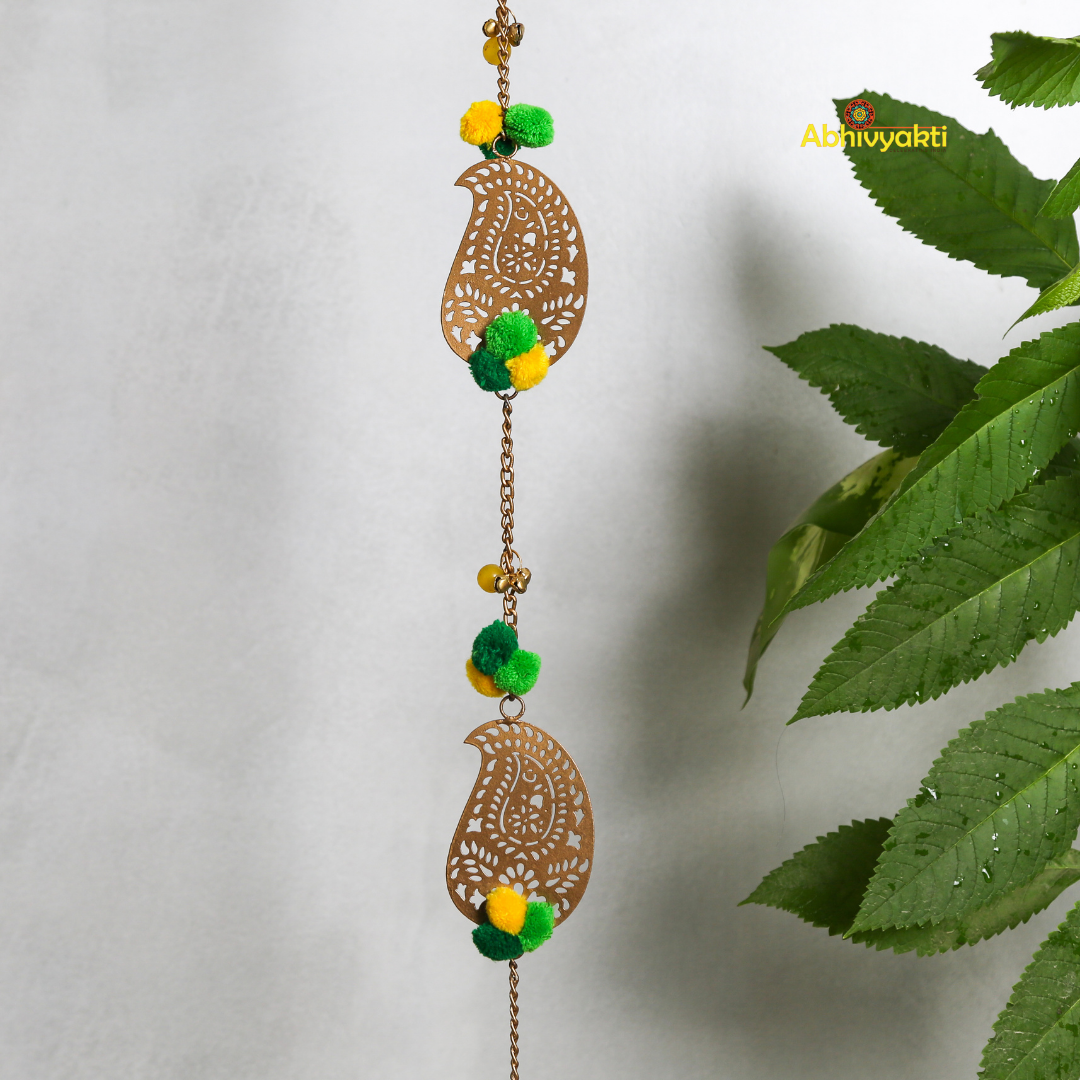 Filigree paisley-shaped metal door hanging adorned with yellow and green pom-poms and tassels, hanging on a white wall near a wooden door