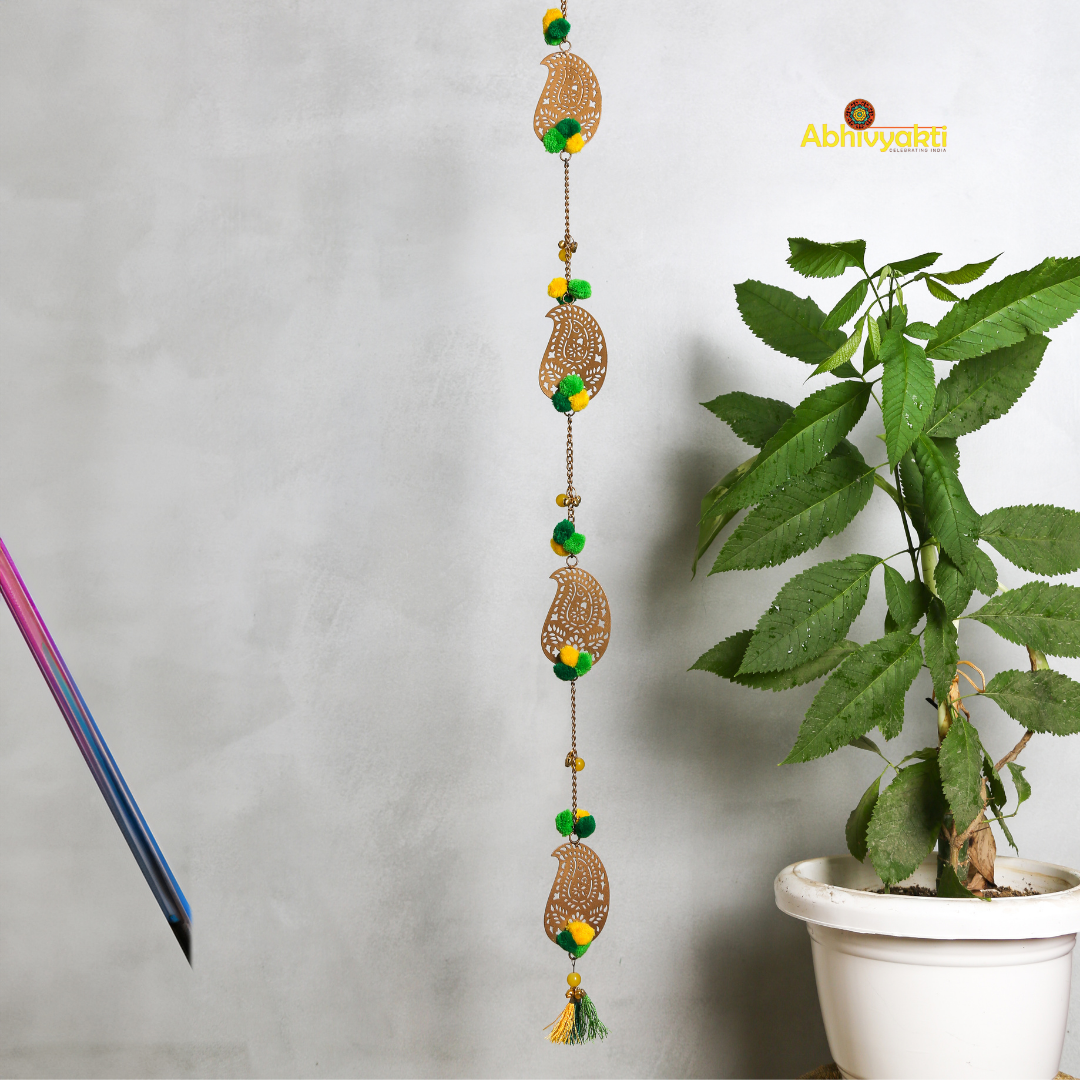 Filigree paisley-shaped metal door hanging adorned with yellow and green pom-poms and tassels, hanging on a white wall near a wooden door