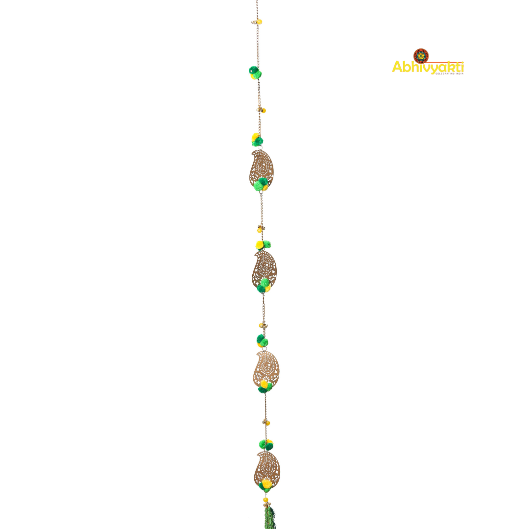 Filigree paisley-shaped metal door hanging adorned with yellow and green pom-poms and tassels, hanging on a white wall near a wooden door