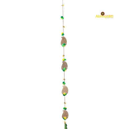 Filigree paisley-shaped metal door hanging adorned with yellow and green pom-poms and tassels, hanging on a white wall near a wooden door