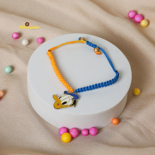 Kids rakhi featuring a Donald Duck cartoon charm on a dual-colored braided thread (orange and blue), displayed on a white round stand with colorful fabric balls scattered on a beige fabric background