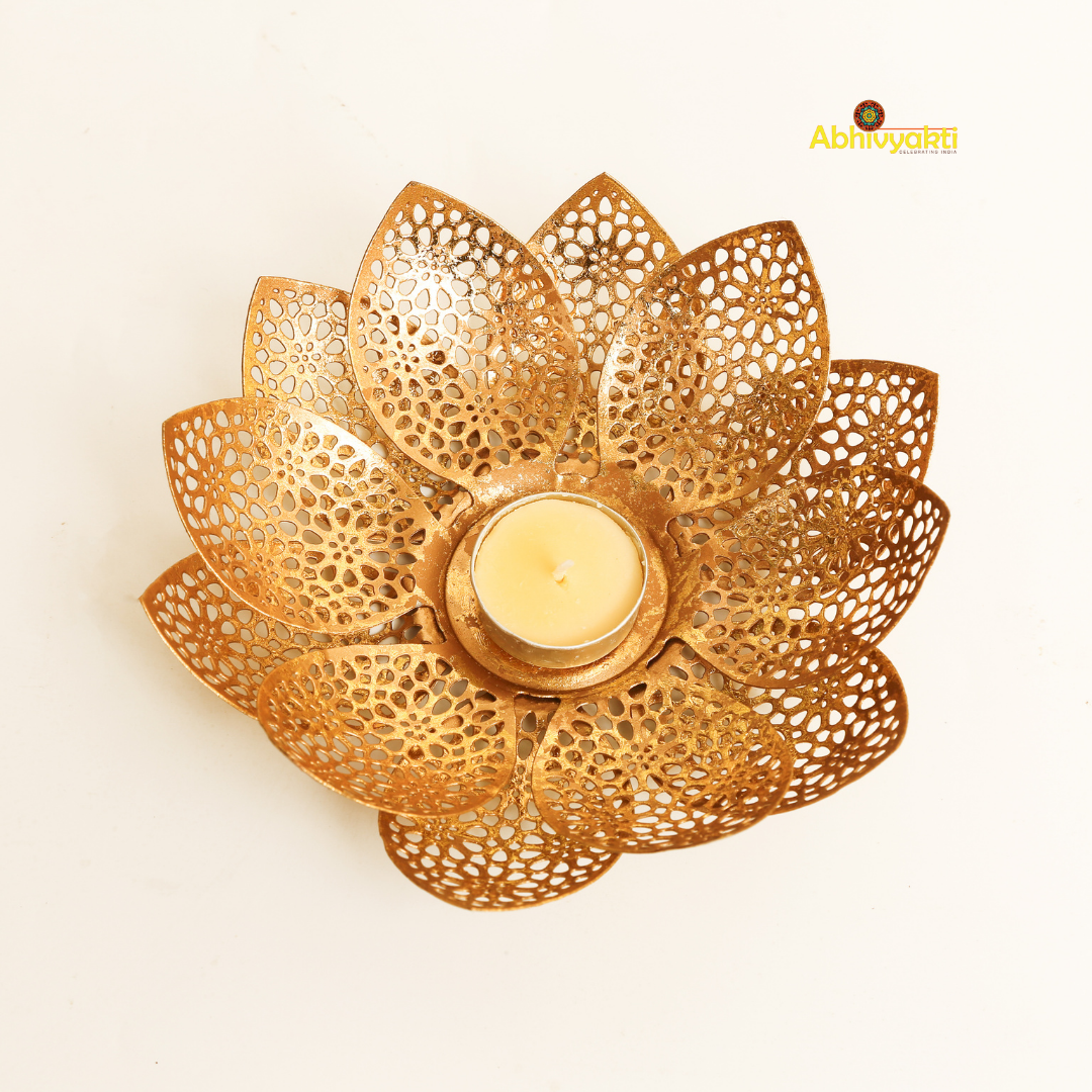 Gold-colored metal lotus flower-shaped t-light holder Padam on a white surface, with a white t-light candle inside, accompanied by artificial yellow and orange flowers to the side.