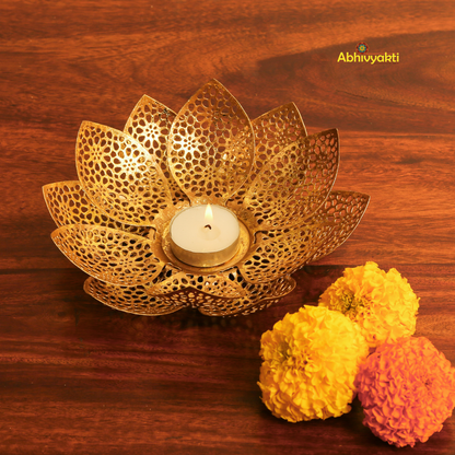 Gold-colored metal lotus flower-shaped t-light holder Padam on a wooden surface, with a white t-light candle inside, accompanied by artificial yellow and orange flowers to the side.