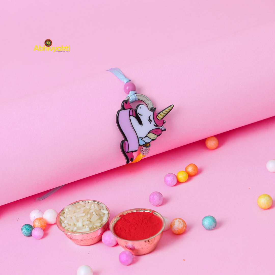 A pink wristband with a cute unicorn charm featuring a pastel-colored mane and horn. The unicorn, smiling with closed eyes, is adorned with a decorative banner. Perfect as a rakhi for kids, the wristband also has a yellow ribbon and metallic spring detail against its pink background.