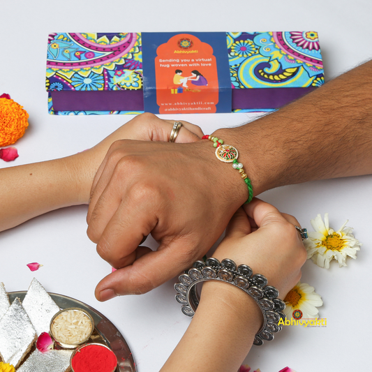 An ornate premium rakhi with a braided red and green thread and golden beads. The center pendant features a colorful, intricate multicolor floral kundan design with green, red, and pearl-like details against a light blue background.