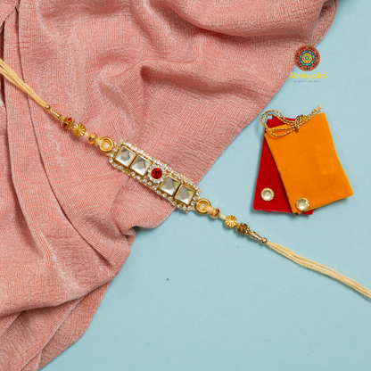 Traditional Rakhi with a rectangular gold centerpiece featuring three stones and a red stone in the center, decorated with gold beads and cream threads, placed on a light blue background with a pink cloth and two small pouches