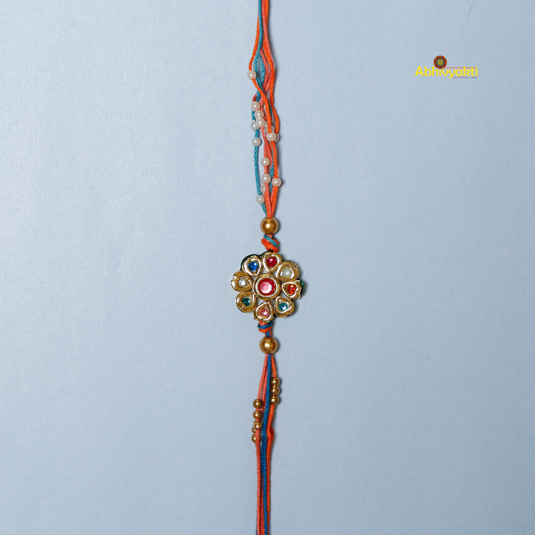 Traditional Rakhi with a round, floral gold centerpiece decorated with multicolored stones and pearls, on orange and blue threads, displayed on a light blue background