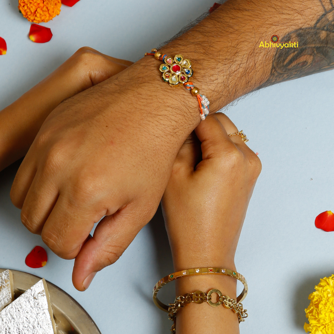 Traditional Rakhi with a round, floral gold centerpiece decorated with multicolored stones and pearls, on orange and blue threads, displayed on a light blue background