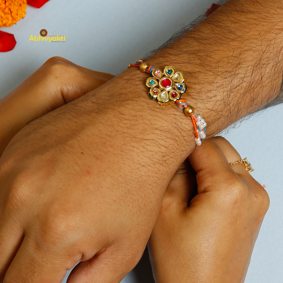 Traditional Rakhi with a round, floral gold centerpiece decorated with multicolored stones and pearls, on orange and blue threads, displayed on a light blue background