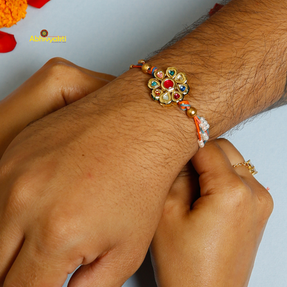Traditional Rakhi with a round, floral gold centerpiece decorated with multicolored stones and pearls, on orange and blue threads, displayed on a light blue background
