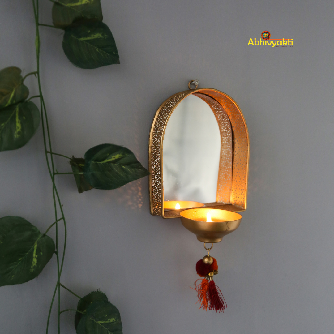 A wall-mounted brass door mirror with a decorative border and a red hanging ornament, displayed against a grey background with green leaves to the side.