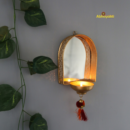 A wall-mounted brass door mirror with a decorative border and a red hanging ornament, displayed against a grey background with green leaves to the side.