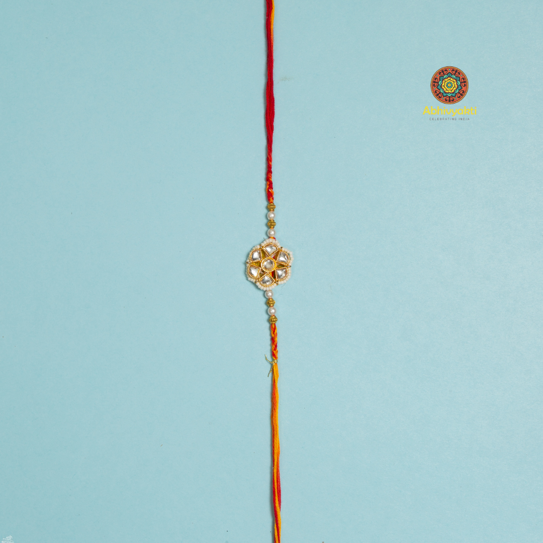 Elegant designer rakhi featuring a floral-shaped golden centerpiece adorned with pearls and stones, laid on a rolled blue background with decorative pearls, rice, and red kumkum in small golden bowl