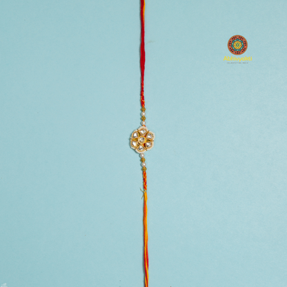 Elegant designer rakhi featuring a floral-shaped golden centerpiece adorned with pearls and stones, laid on a rolled blue background with decorative pearls, rice, and red kumkum in small golden bowl