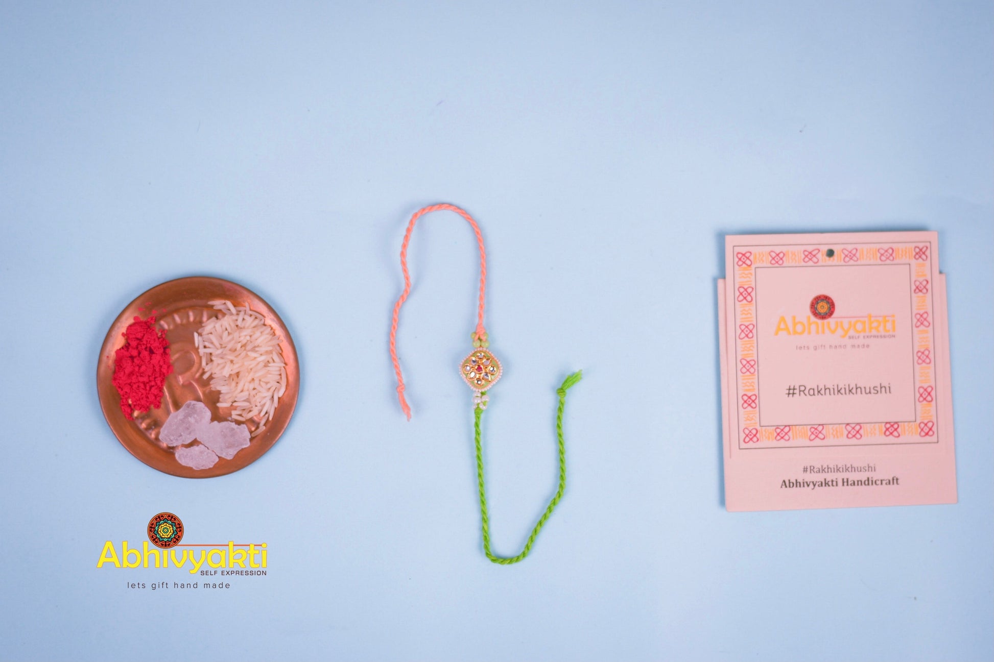 Elegant rakhi with a gold and green centerpiece, decorated with small pearls and colored stones, tied with green and peach threads, placed on a blue background with rice and kumkum