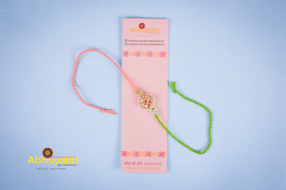 Elegant rakhi with a gold and green centerpiece, decorated with small pearls and colored stones, tied with green and peach threads, placed on a blue background