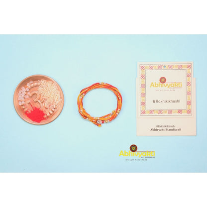 Simple stone rakhis with red and yellow threads and small gold beads, one with a clear stone in the center, all wrapped around a blue roll. Bowls of rice and roli, scattered pearls